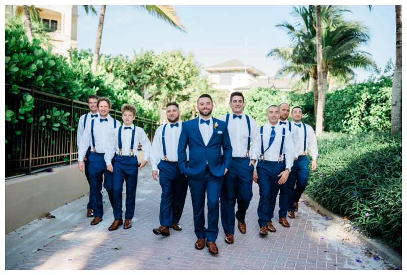 Groom and groomsmen dressed in a navy suit and navy suspenders walk smiling towards wedding ceremony.