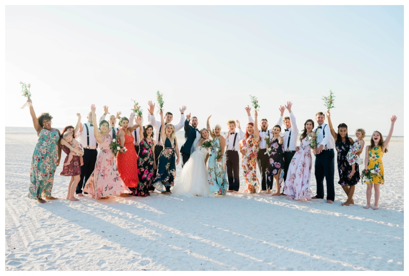 Large group of bride, groom and bridal party excitedly cheer on the beach after wedding ceremony.