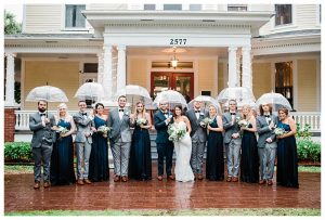 Bridal party stands under clear umbrellas on rainy wedding day in front of historic Heitman House wedding venue