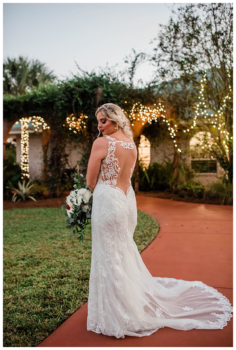 Bride shows details of backless wedding gown under fairy lights.