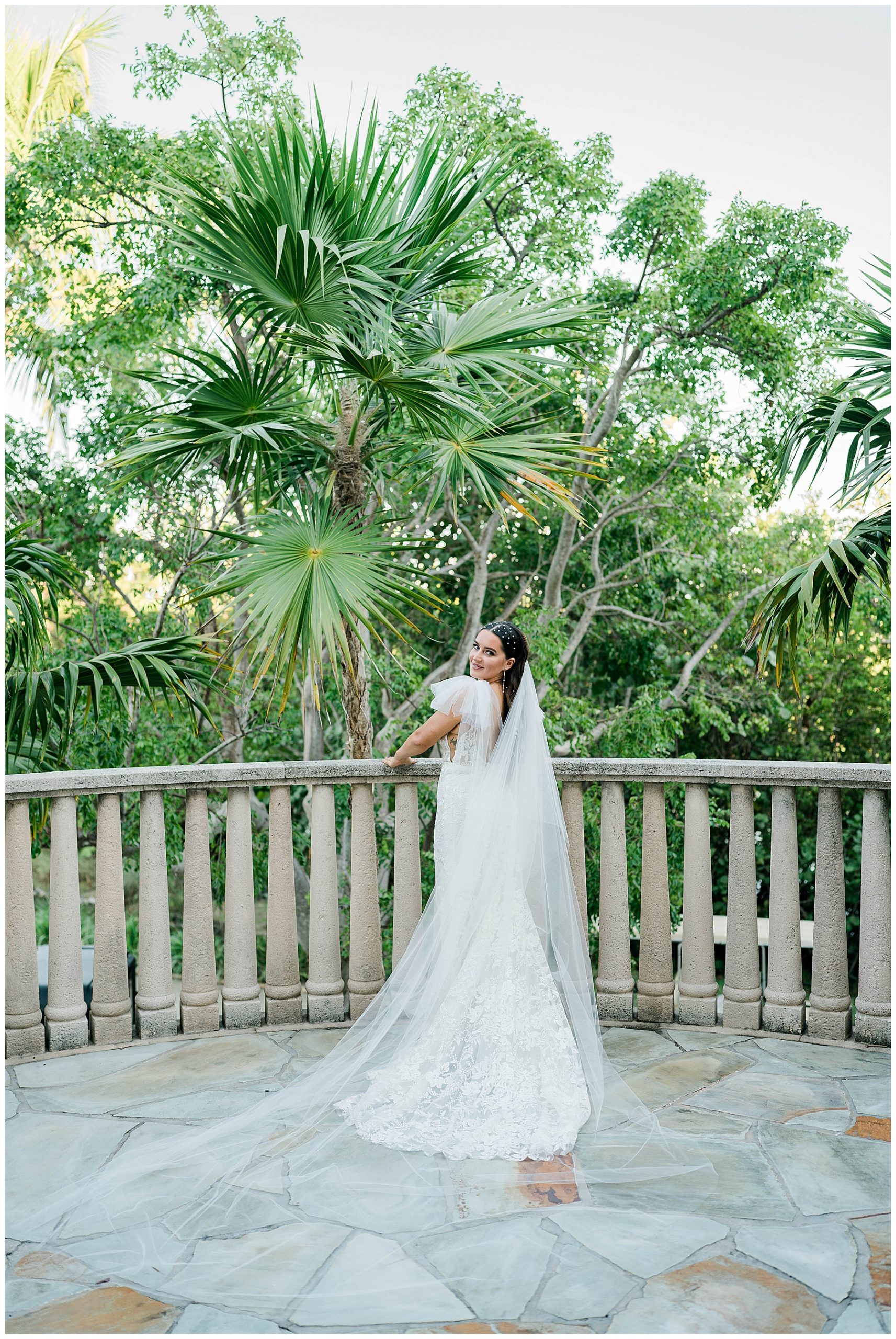 bride stands on stone balcony in long bridal gown and cathedral bridal veil during Southwest Florida wedding day