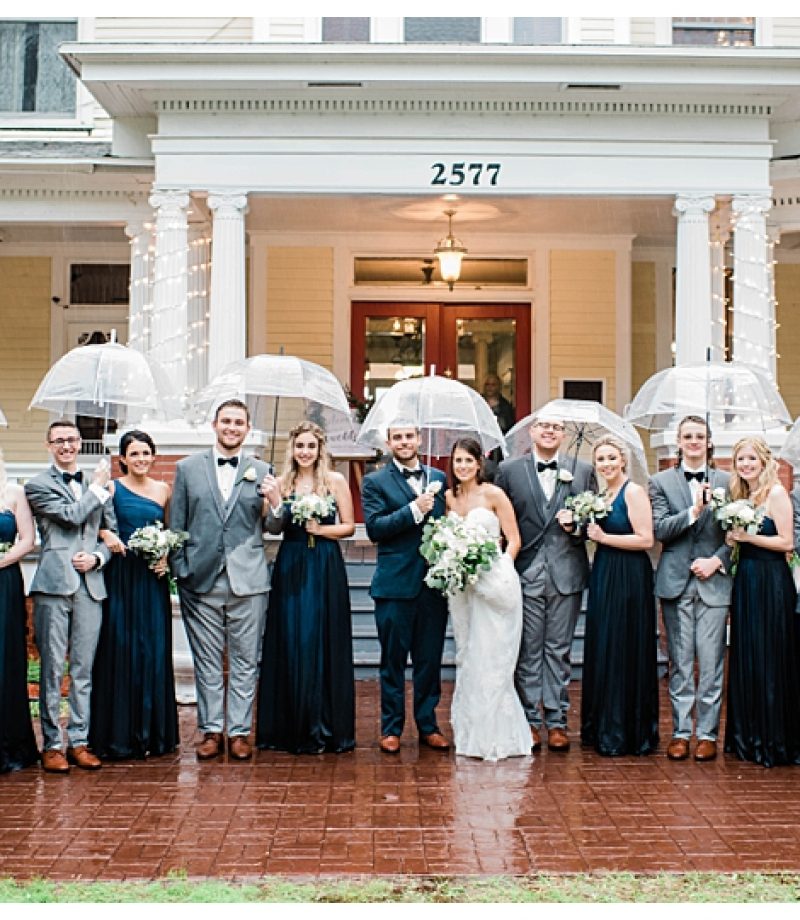 Bridal party stands under clear umbrellas on rainy wedding day in front of historic Heitman House wedding venue