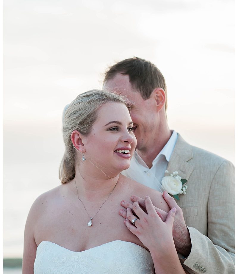 Bride smiles out to the sunset as groom kisses her on Southwest Florida beach wedding day